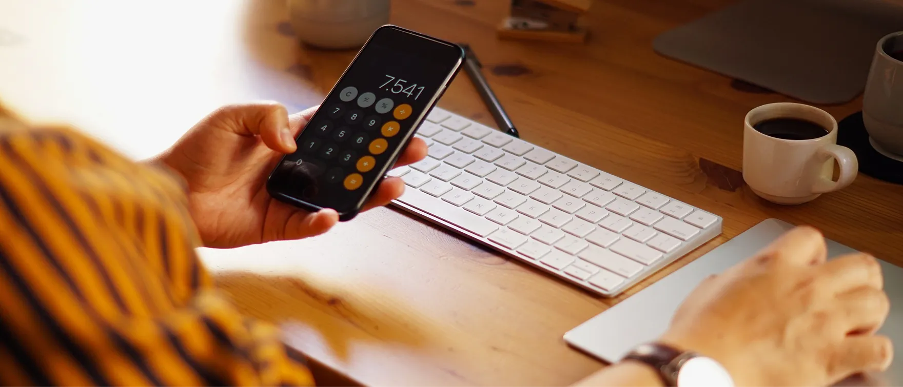 Une main tenant un téléphone avec une calculatrice, clavier et tasse de café en arrière-plan.