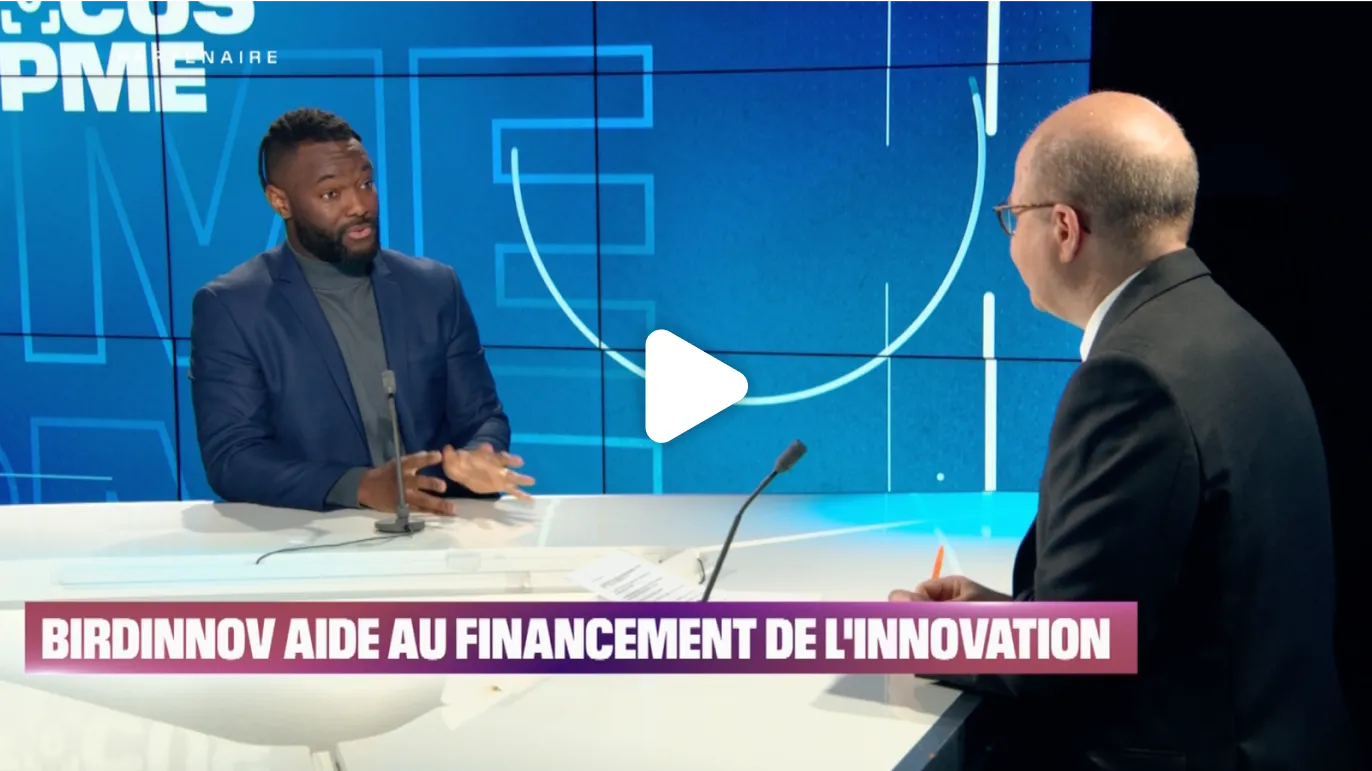 Deux hommes discutent sur un plateau, avec des graphiques en arrière-plan.