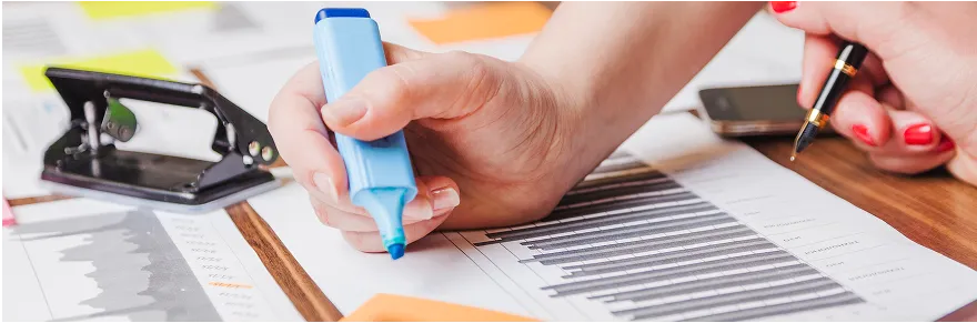 Main tenant un surligneur bleu sur des documents avec divers outils de bureau autour.