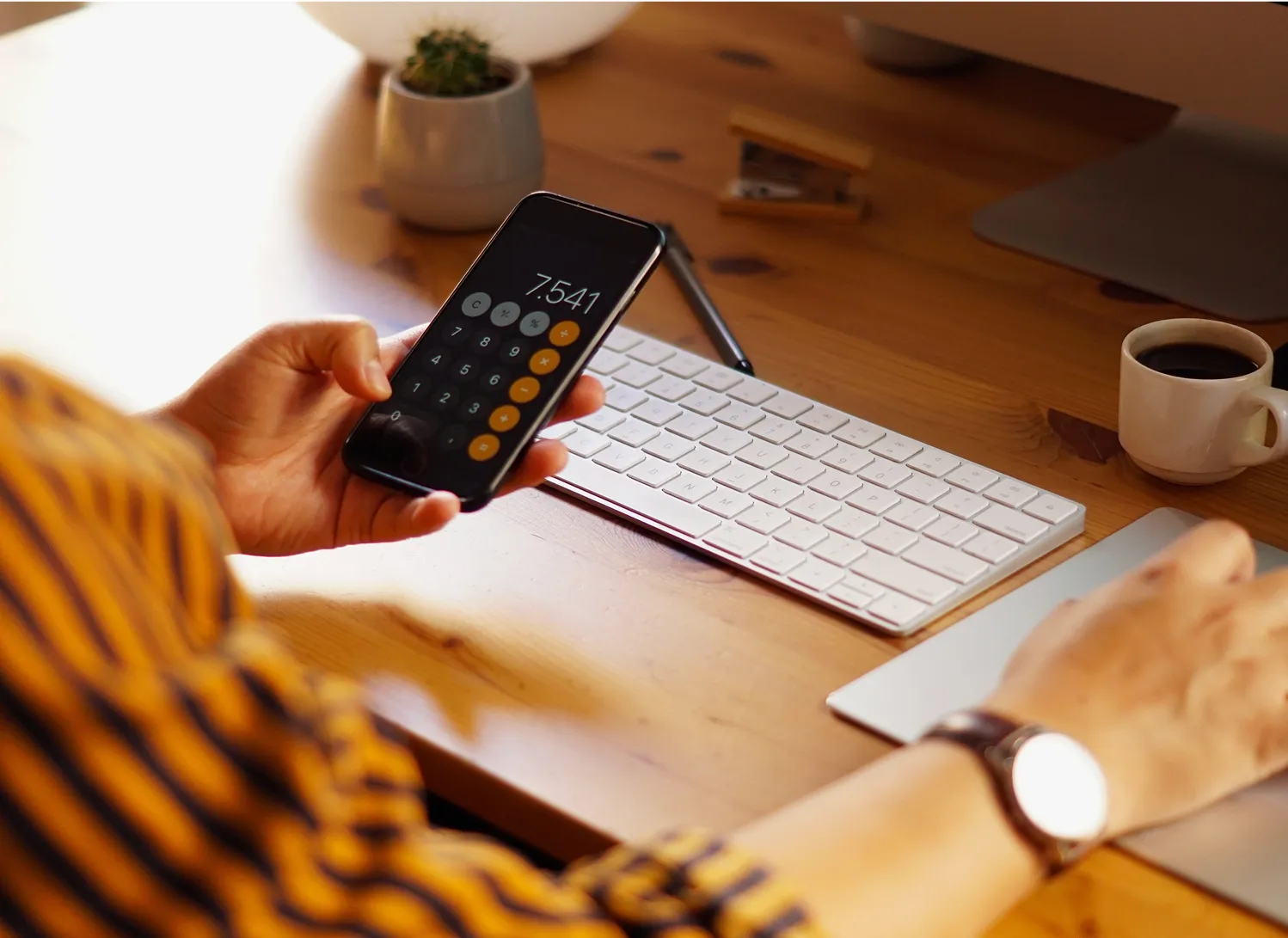 Une main tenant un smartphone avec une calculatrice ouverte sur un bureau en bois.