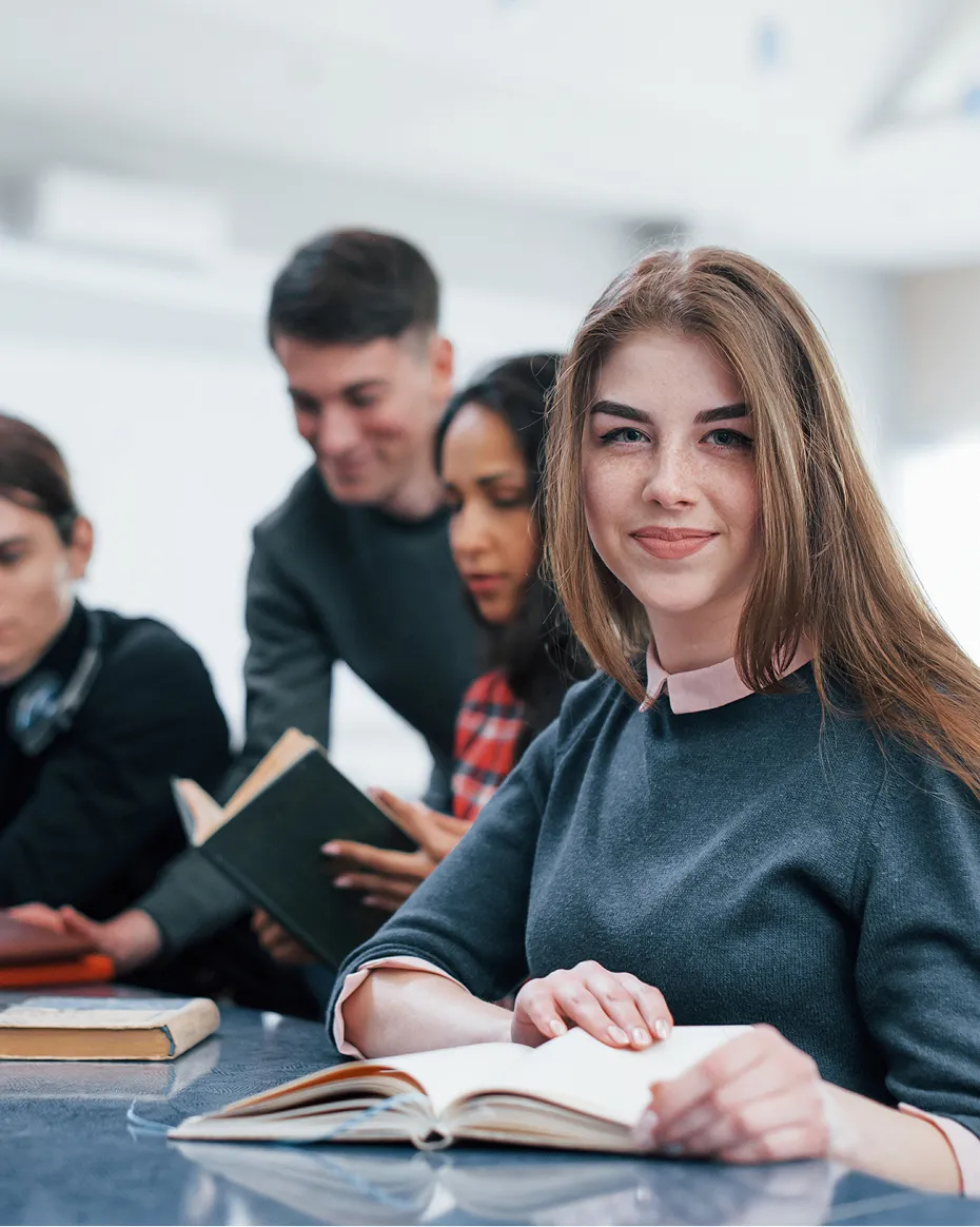 Étudiante souriante, un livre ouvert devant elle, entourée de camarades en discussion.