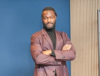 Homme souriant, bras croisés, vêtu d'un costume marron, devant un mur bleu.